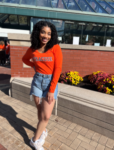 Photo of Chesnie Caster outside in the sun standing in front of a building. She is wearing an orange Syracuse shirt and denim shorts.